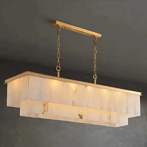 An elegant rectangular brass chandelier with a gold frame and chain hangs from the ceiling. The light fixture boasts an alabaster-like texture with a layered, frosted glass design, emitting a warm, soft glow. This striking Shopp578 Natural Marble Dining Room Chandelier contrasts beautifully against the simple, dark gray wall.