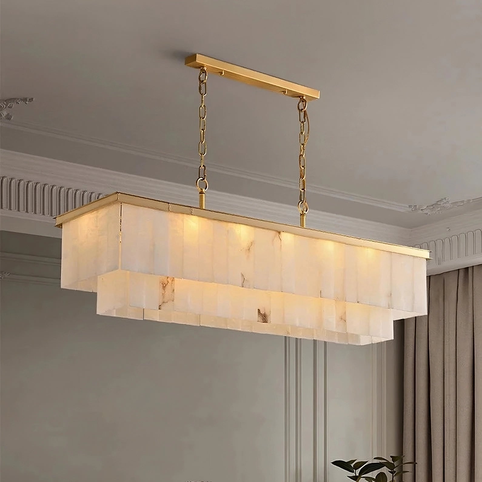 A **Shopp578 Natural Marble Dining Room Chandelier** with a gold frame and brass accents hangs from the ceiling by two gold chains. The chandelier features multiple layers of translucent panels, softly glowing with warm light, illuminating the elegant room with decorative molding and beige curtains.