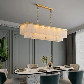 A modern dining room features a rectangular marble table with dark green chairs arranged around it. Above the table hangs a Shopp578 Natural Marble Dining Room Chandelier with a gold frame and layered design. The room has light grey paneled walls, a large window with grey curtains, and exudes sophistication with its natural marble elements.