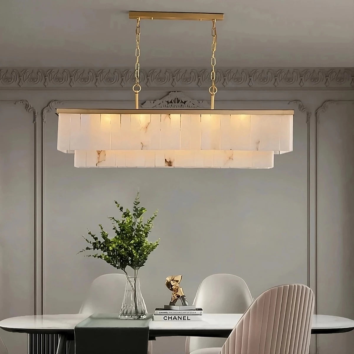 A dining room features a modern brass chandelier, the Shopp578 Natural Calcite Dining Room Chandelier, with two tiers of rectangular natural calcite panels, emitting a warm light. Below, a white dining table holds a clear vase with green foliage and a small decorative item. The chairs are neutral tones, and the walls are gray.