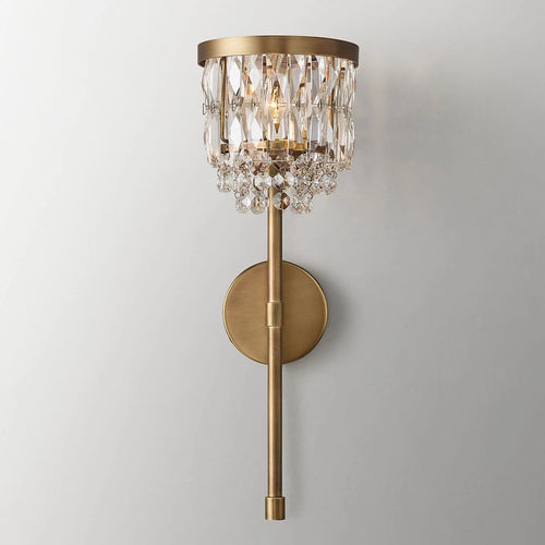 The Valente Crystal Wall Sconce by Morsale.com features a solid brass round base, vertical rod, and hanging crystal prisms for a touch of modern elegance on any light gray wall.