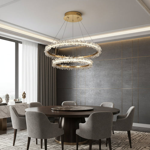 Modern dining room with a round dark wood table, six gray chairs, and the Capri 2-Ring Modern Crystal Chandelier by Morsale.com. Gray walls, gold accents, and a large window elevate the space’s modern luxury lighting and natural ambiance.