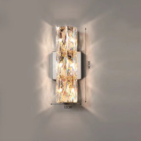 The Bacci Crystal Wall Light Sconce by Morsale.com is a modern indoor fixture boasting a sleek chrome base and a rectangular design with layered, transparent crystal accents. The lamp measures 12 cm in width and 30 cm in height, casting a warm, ambient glow on the gray wall behind it.