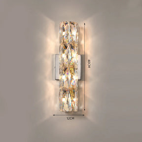 The Bacci Crystal Wall Light Sconce from Morsale.com emits a warm light. This handmade fixture is rectangular and features multiple faceted crystal pieces, measuring 40 cm in height and 12 cm in width. The LED dimmable crystals refract light, creating a sparkling effect on the surrounding wall.