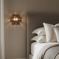 A modern bedroom corner featuring a padded headboard, white pillows, beige bedding, and the Riviera Smoke Glass Sconce from Morsale.com casting soft, warm light on a neutral wall for luxurious interior lighting.