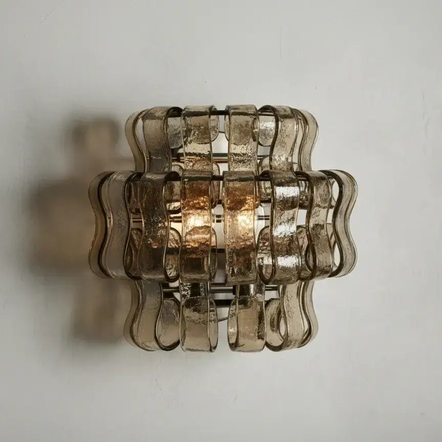 The Riviera Sconce by Morsale.com features a sculptural curved glass design with textured wavy strips, forming a geometric square. It emits a warm glow and soft shadows, perfect for adding luxurious lighting to any space.