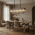 Elegant dining room featuring a wooden table, six beige upholstered chairs, the Gio Crystal Dining Chandelier by Morsale.com above, large window with sheer curtains, decorative vases, and a white orchid centerpiece.