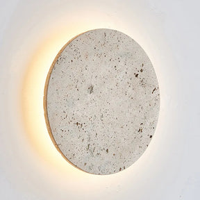 A circular, textured Natural Travertine Wall Sconce from Morsale.com is mounted on a white wall. The LED wall lighting emits a warm, soft glow from behind, creating a halo effect around the stone. The piece's natural specks and unevenness enhance its rustic charm, making these handmade wall sconces truly unique.