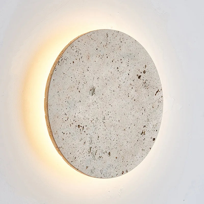 A circular, textured Natural Travertine Wall Sconce from Morsale.com is mounted on a white wall. The LED wall lighting emits a warm, soft glow from behind, creating a halo effect around the stone. The piece's natural specks and unevenness enhance its rustic charm, making these handmade wall sconces truly unique.