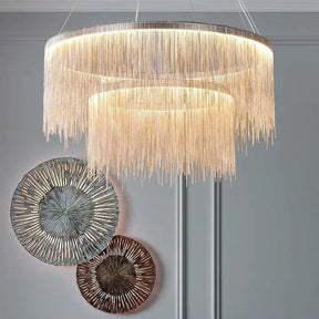 The Cadena Steel Tassel Modern Chandelier by Morsale.com, featuring multiple layers of cascading thin golden chains in a large circular design, serves as the focal point with its sleek LED light source. The backdrop of marble walls with a white and gray veined pattern underscores the elegance of the modern interior design.