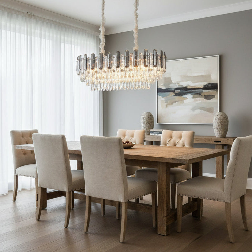 A modern dining room features a large wooden table, six upholstered chairs, neutral walls, abstract art, two vases on a sideboard by a window with sheer white curtains, and a Venice Crystal Dining Room Chandelier from Morsale.com.