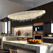 A modern kitchen with a large, oval Alleri Crystal Chandelier For Dining Room by Morsale.com made of handmade premium crystals hanging above a marble island. The island features a fruit bowl filled with apples. The kitchen cabinets are dark wood, with built-in stainless steel appliances and a marble backsplash.