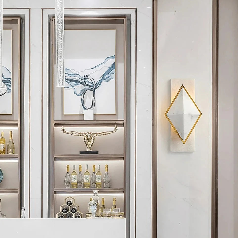 A modern, sleek interior featuring a white countertop made of Natural Spanish marble in the foreground with bottles displayed behind it. The wall behind showcases several gold and silver decorative items, shelves with more bottles, a Bigman Natural Marble Contemporary Wall Sconce Light, and framed art with abstract designs.