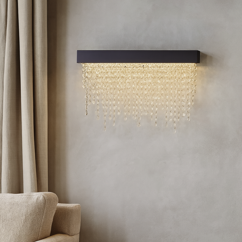 
                  
                    The Alleri Crystal Wall Sconce by Morsale.com features a rectangular black base and cascading hand-cut crystal-like beads, shown mounted on a gray wall beside beige curtains and a light upholstered chair.
                  
                