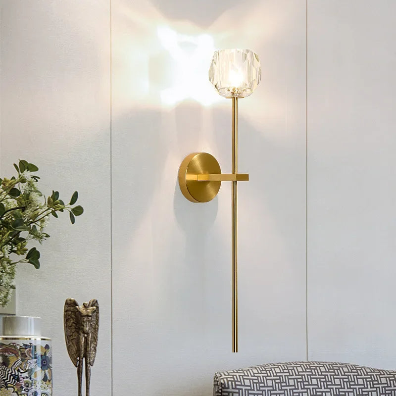 A stylish and sophisticated Clear Crystal & Brass Wall Light Fixture from Morsale.com provides ambient lighting on a light-colored wall. Below the fixture, a small plant and metallic decorative figure sit gracefully, while the corner of a patterned piece of furniture is visible at the bottom right.