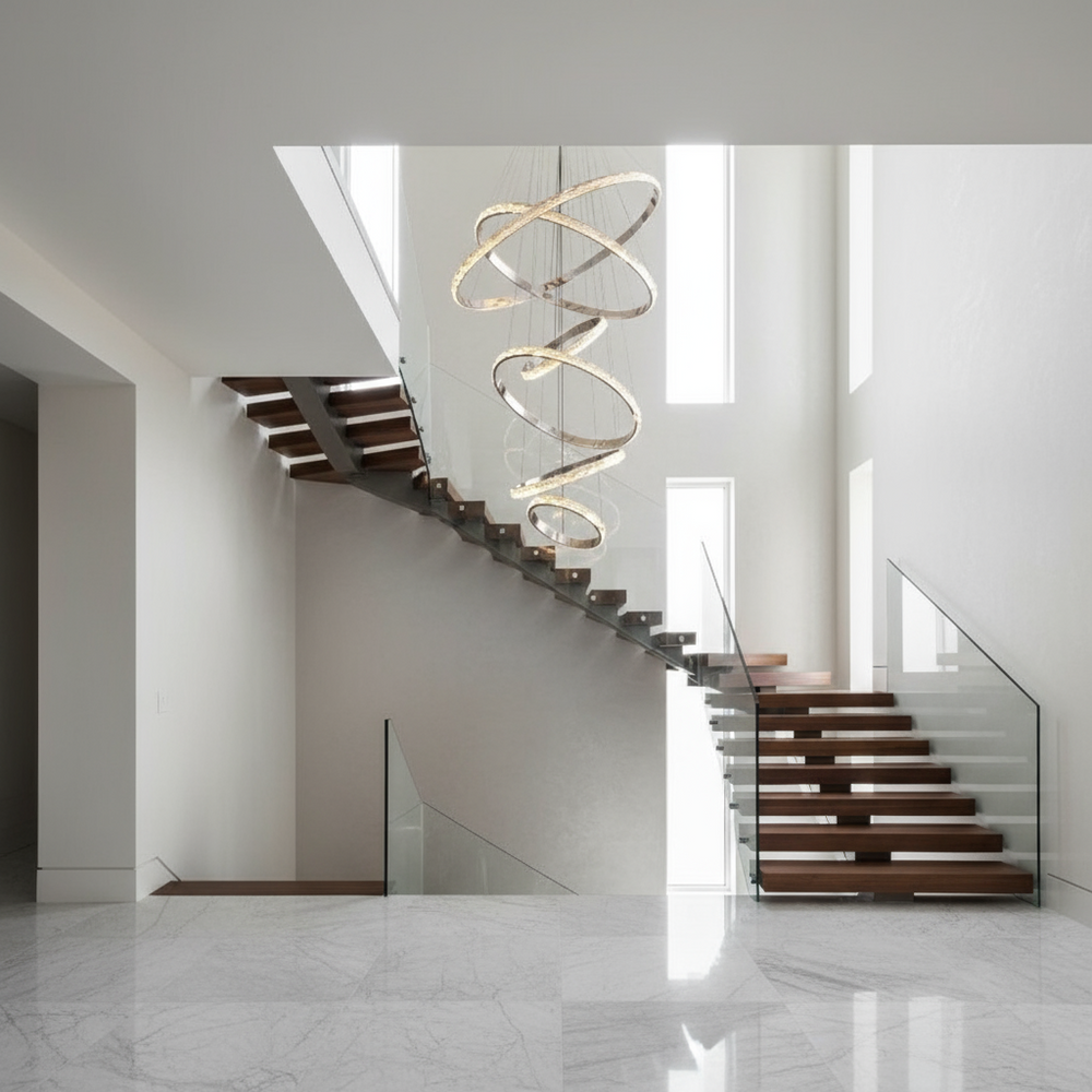 A modern staircase with dark wooden steps and glass railings rises beside tall windows. Above, the Argento 6-Tier Crystal Ring Chandelier from Morsale.com shines, while the floor features polished white marble with subtle gray veining.