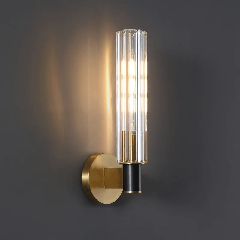 The Morsale Palermo Retro Brass Wall Sconce features a brass finish with a cylindrical glass shade, mounted on a circular base against a dark gray wall. The light is turned on, emitting a warm glow.