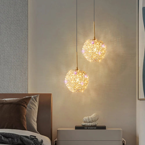 Two Amri Crystal Pendants from Morsale.com hang above a modern nightstand beside a bed with neutral bedding, softly illuminating the wall. A decorative white object rests on the nightstand, completing the elegant look.