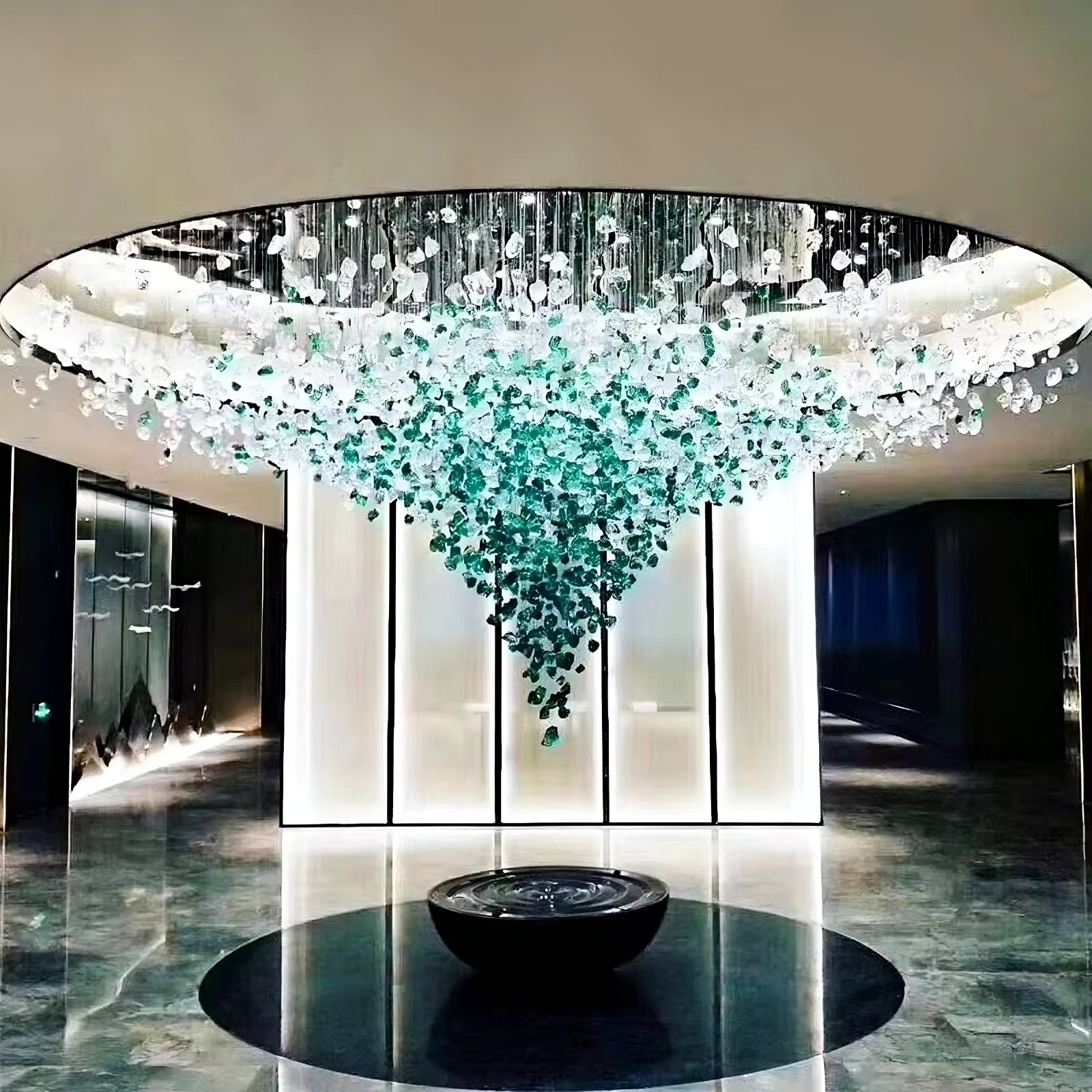 A modern lobby features the Morsale.com Piovere Multi Pendant Chandelier, resembling a crystal chandelier with cascading green and clear glass crystals. Below it, a round black planter rests on the polished floor, complemented by modern lighting that accentuates the sleek illuminated wall.