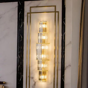 The Gio 32" Crystal Sconce by Morsale.com enhances a marbled wall with its modern design, featuring vertical gold and glass elements that emit a warm, ambient glow. Framed by dark and light marble patterns, this sconce contributes to a luxurious atmosphere with its sophisticated aesthetic.