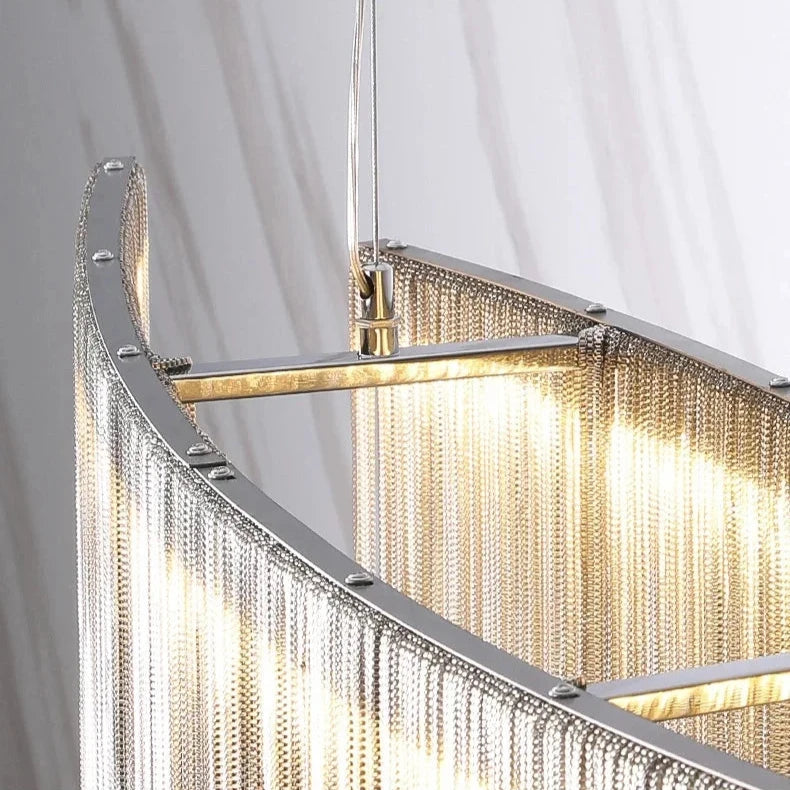 A close-up view of the sleek, modern Cadena Stainless Steel Dining Room Light Fixture by Morsale.com. Its stainless steel frame is adorned with cascading chain decorations, and the LED light source illuminates the fixture, highlighting its intricate design and the fine details of the chains. The background features a neutral-colored surface.