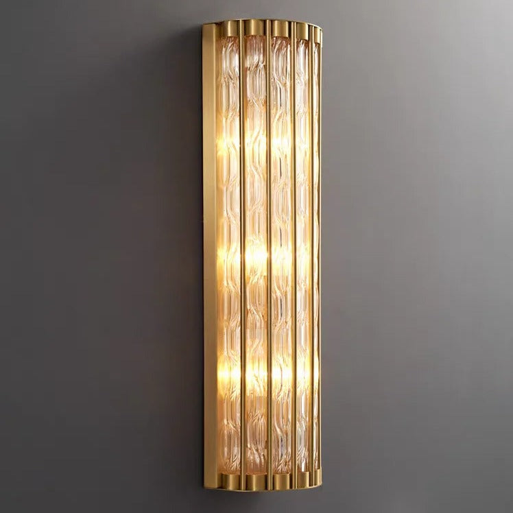 A vertically mounted, rectangular Modern Copper & Glass Wall Sconce from Morsale.com features parallel glass rods within a brass frame, with internal lights casting a warm, inviting glow. The textured glass design adds an elegant touch to the modern fixture, which is set against a plain gray wall.