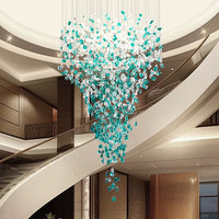 The Morsale Verde Crystal Foyer Chandelier, featuring cascading clear and teal glass, hangs in the center of a modern lobby with curved staircases and neutral walls, embodying the essence of contemporary lighting.
