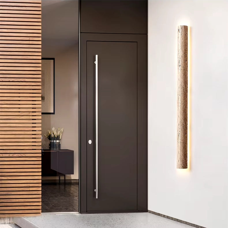 A modern entrance showcases a tall, sleek black door with a long silver handle. On the adjacent white wall, a vertical fixture called the IP65 Waterproof Natural Travertine Wall Light from YEBMLP, featuring sustainable lighting, is mounted. To the left, part of a wooden slatted partition can be seen. A plant adds decorative charm to the interior.