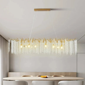 A modern, rectangular Albero Kitchen Ceiling Light Fixture from Morsale.com hangs above a dining table, featuring cascading crystal-like elements and multiple light bulbs. The room has a minimalist design with neutral tones, and a bowl of fruit can be seen on the table below the chandelier.