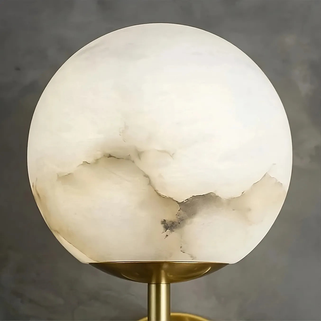 The Natural Marble Sphere Wall Sconce from Morsale.com, with unique sedimentary patterns in shades of white and gray, is mounted on a brass-colored stand, casting a warm glow against a soft gray background.