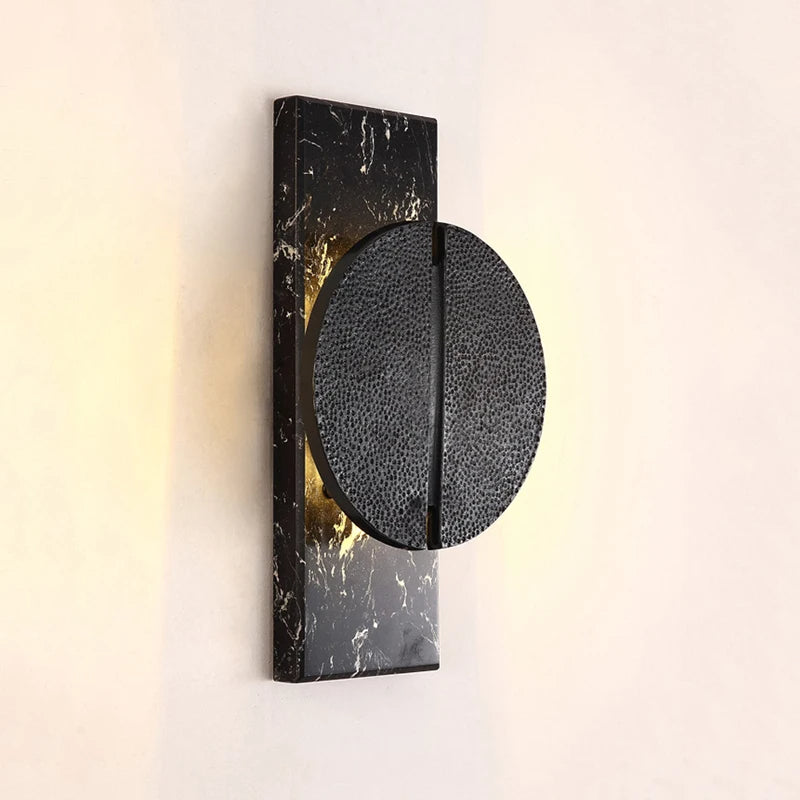 A modern Bigman Medieval Marble Wall Light Sconce features a textured black circular element affixed to a rectangular base of natural Spanish marble. The light glows softly around the edges, highlighting the textures and art deco glamor against a plain tan wall.
