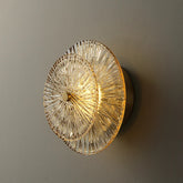 A Modern Glass Wall Sconce from Morsale.com with a round, intricate glass design illuminates a gray wall. The glass features a radial pattern and emits a warm, golden light that adds a touch of elegance to the space. This handmade wall sconce combines artistry with functionality, perfect for any sophisticated setting.