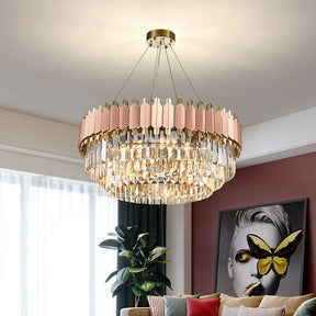 A modern living room featuring a Gio Rose Gold Modern Chandelier by Morsale.com with gold-plated stainless steel and pink accents hanging from the ceiling. The room has a brown accent wall with an artistic portrait of a woman's face adorned with a butterfly. A potted plant and sofa are also visible.