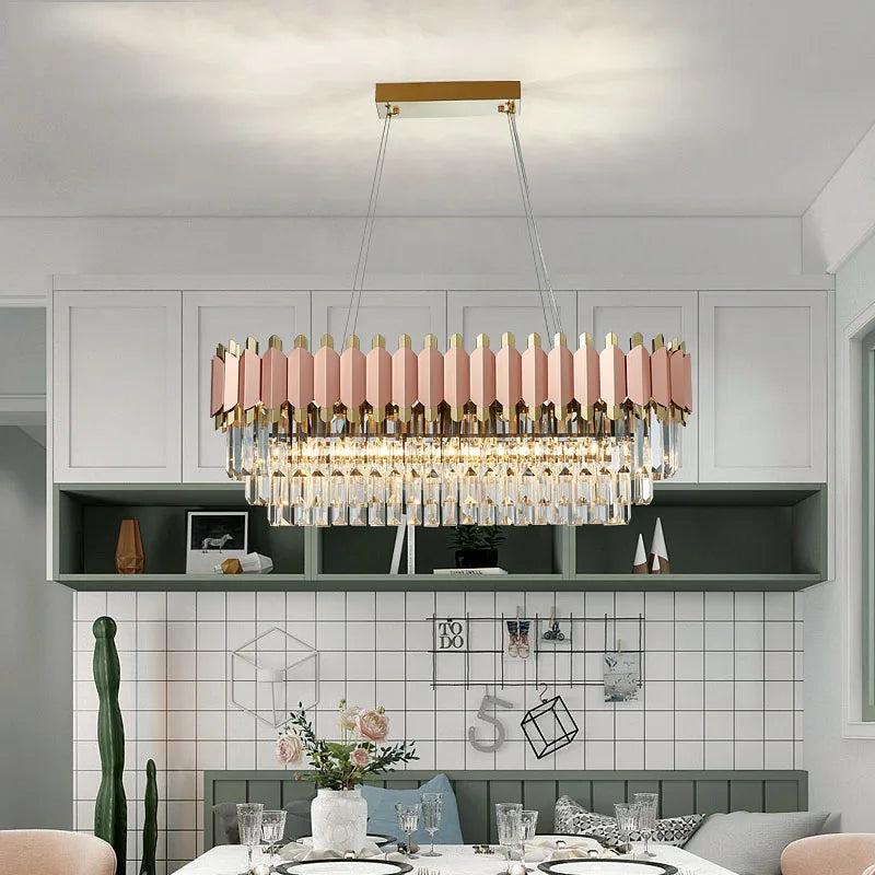 A modern dining room features a rectangular glass table set with plates and glasses. Above the table, the Gio 39" Rose Gold Dining Room Chandelier from Morsale.com dazzles with its mix of pastel pink and gold segments and handmade crystals. The background boasts white cabinets and green shelving, enhancing the elegant ambiance.