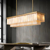 A modern dining room showcases the striking Moonshade Marble Dining Room Chandelier by Shopp578, featuring layered, translucent beige panels. The chandelier hangs elegantly above a wooden table with cushioned chairs. The wall is a subtle gray, and natural light streams in from a window on the left.