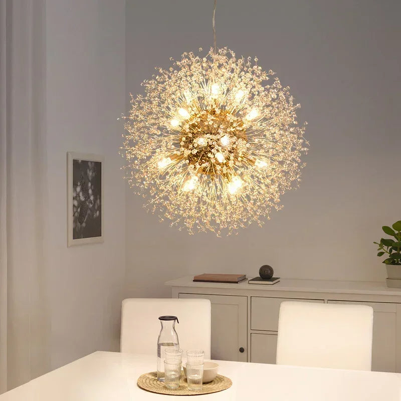 A modern dining room features a round table, white chairs, and the Morsale.com Dandelion Crystal Globe Chandelier glowing with LED bulbs. A white cabinet, potted plant, and framed art complete the background.