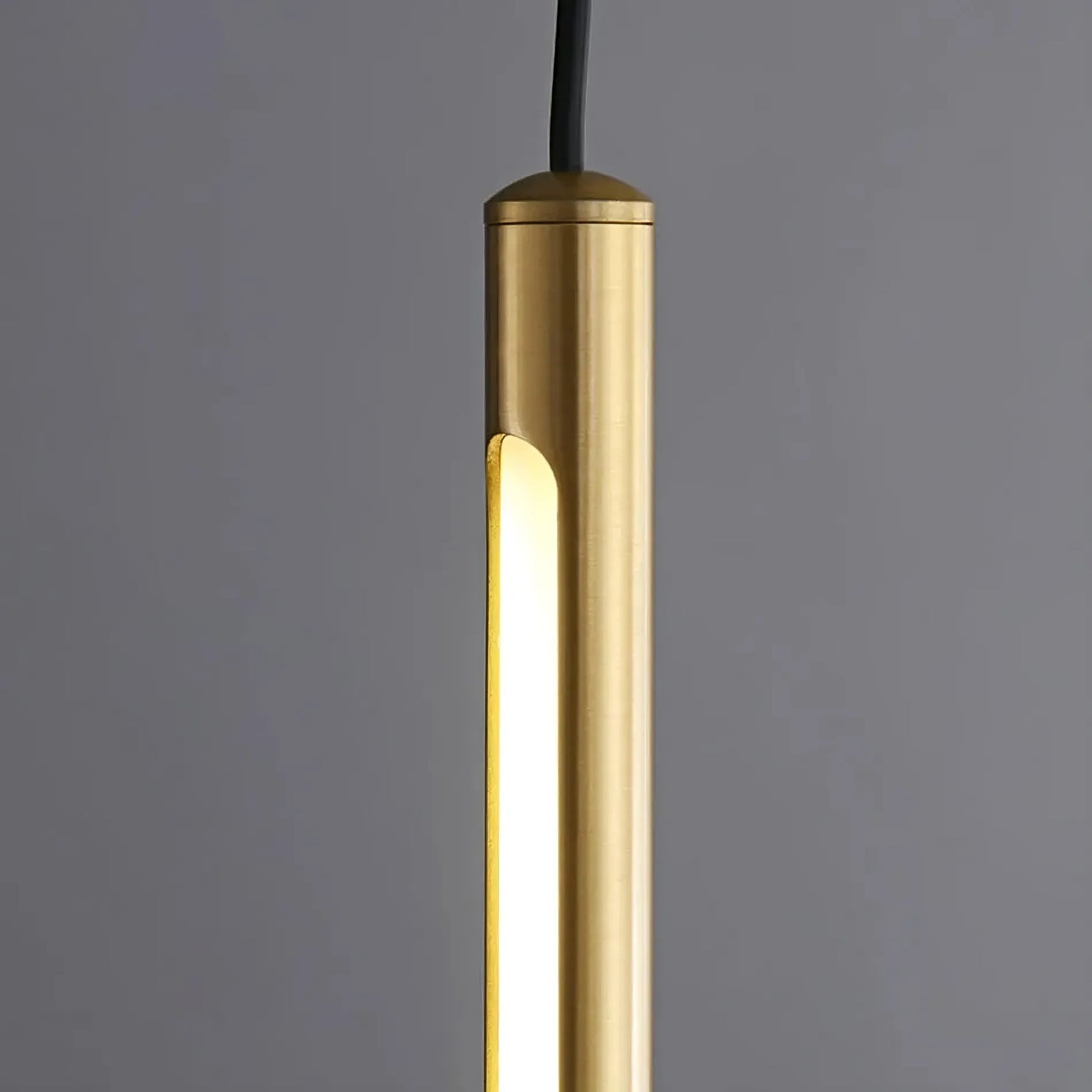 The Grande Brass Entryway Pendant Light Fixture by Morsale.com is depicted suspended against a gray background. This cylindrical pendant light features a brushed brass finish and emits a warm, diffused glow from an elongated vertical opening on its side, enhanced by modern LED chips. A dimmable design adds versatility, while a black cord is visible at the top leading to the ceiling.