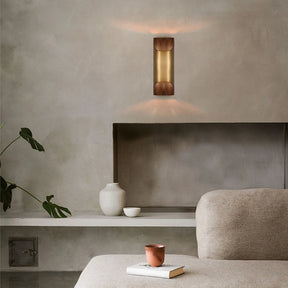 A minimalist living room includes a beige couch, a small table holding a red cup, and a decorative shelf showcasing white vases. The Morsale Rame Modern Copper Wall Lights cast light both upwards and downwards against a textured wall, enhancing the modern, serene atmosphere with elegant home decor.