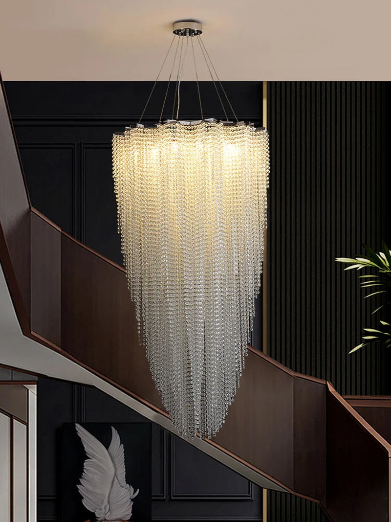 An elegant, luxurious Alleri Crystal Entryway Chandelier from Morsale.com with cascading dazzling crystals hangs from the ceiling, illuminating a modern interior. The chandelier's crystals sparkle, adding a touch of luxury to the space. Below, a staircase with dark wooden railings is visible, and there is a decorative framed artwork of a white feather on the wall.