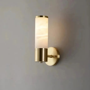 The Moonshade Natural Marble Sconce Light from Morsale.com, featuring a modern design with a cylindrical frosted glass shade and brushed gold hardware, is mounted on a plain grey wall. The fixture, compatible with an E14 base LED, is turned on, emitting a warm glow.