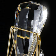 A detailed close-up of a faceted, clear crystal held in place by a sleek, geometric gold frame. The crystal captures and refracts light, creating a subtle spectrum of colors. The background is black, emphasizing the brilliance and clarity of the Villa 8-Light Dining Room Light Fixture by Bigman.