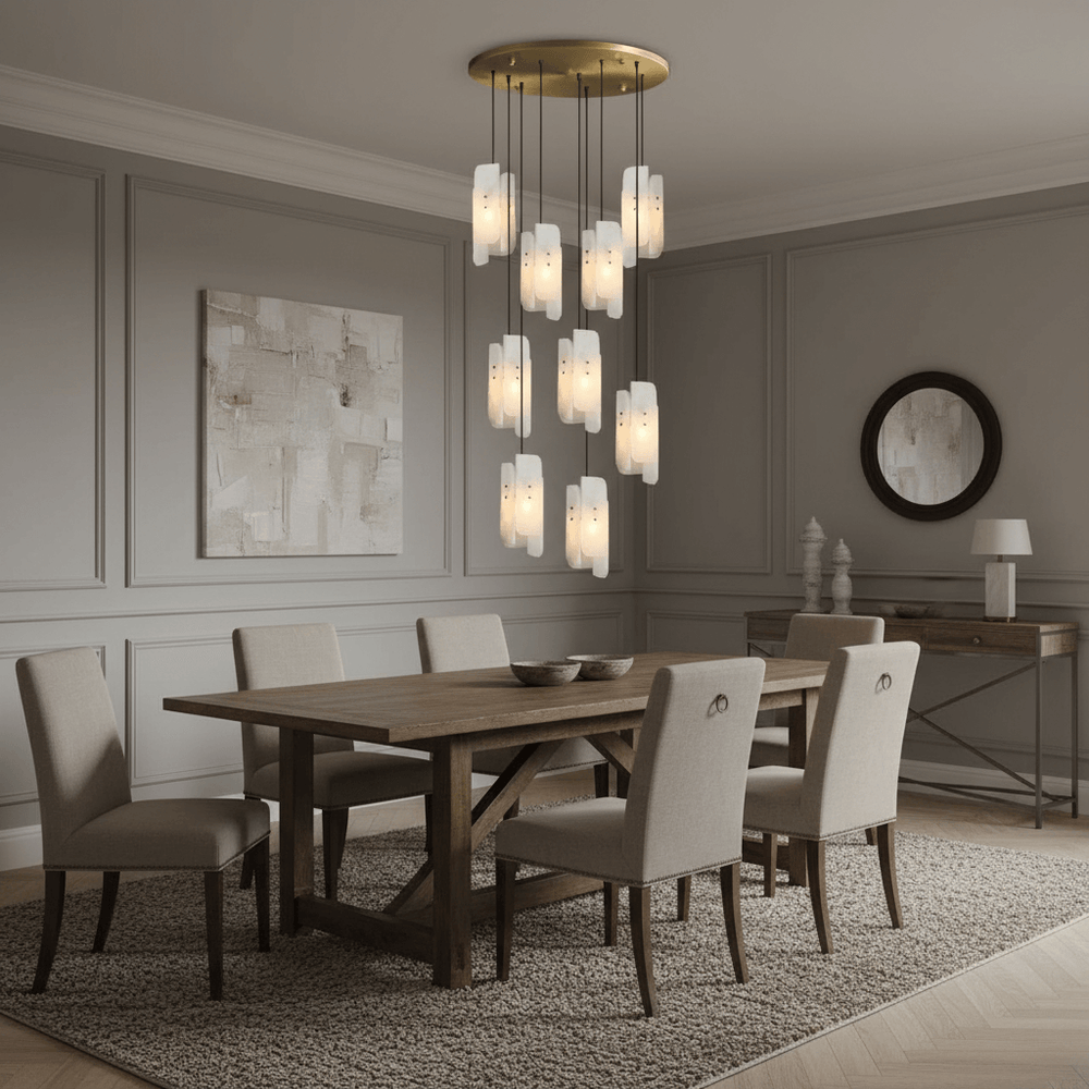 A modern dining room features a wooden table, six beige upholstered chairs, a beige rug, Morsale’s Lunara Marble Chandelier for luxury lighting, gray paneled walls with abstract art, and a round mirror above the sideboard.