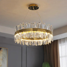 A Morsale.com Calvi Crystal Modern Chandelier with a circular design hangs from the ceiling. It features a gold ring adorned with handmade clear crystals, creating a sparkling light effect. The room has gray walls and a window with cream-colored curtains, allowing natural light to enter.