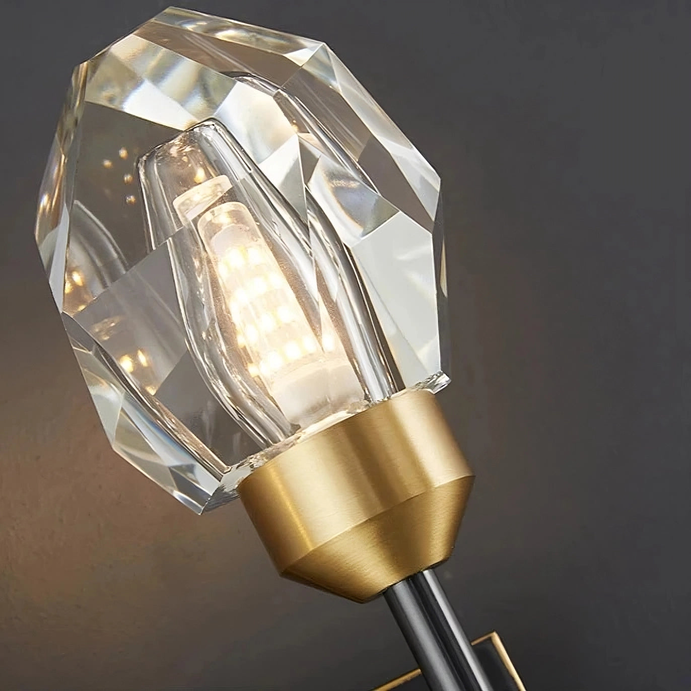 The Morsale Diamante Crystal Wall Sconce, featuring a gemstone-shaped glass shade and a vintage-style LED bulb, is elegantly mounted on a gold-colored base against a dark gray wall. This contemporary lighting design beautifully merges modern and classic elements to provide optimal illumination and a sophisticated appearance.