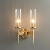 The Morsale Palermo Postmodern Copper Wall Sconce features two cylindrical glass shades and warm glowing bulbs, mounted on a copper-finished base. This piece exemplifies postmodern design and enhances any space with its contemporary elegance when set against a light gray wall.