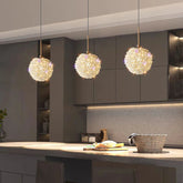 A modern kitchen features sleek dark cabinets and a stone countertop. Three El Sol Crystal Pendant Lights from Morsale.com cast a warm glow. A woven basket and bread-topped cutting board adorn the counter, complementing the stainless steel accents throughout.