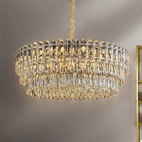 A Gio Crystal Chandelier by Morsale hangs from the ceiling, showcasing multiple tiers of sparkling prisms that beautifully reflect light. Enhanced by energy-efficient LED bulbs against a neutral gray wall, it exudes a modern style and elegant presence.