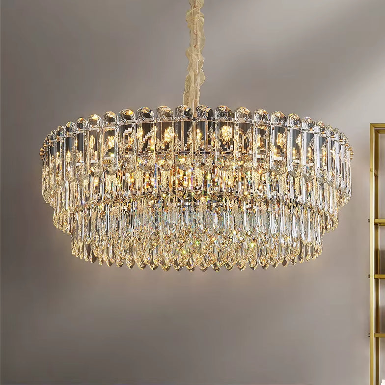 A Gio Crystal Chandelier by Morsale hangs from the ceiling, showcasing multiple tiers of sparkling prisms that beautifully reflect light. Enhanced by energy-efficient LED bulbs against a neutral gray wall, it exudes a modern style and elegant presence.