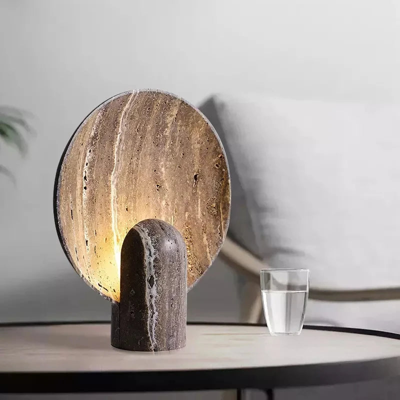 A modern **Morsale Natural Travertine Table Lamp** with a stone-like base and a circular, textured shade that emits a warm light is placed on a round coffee table. Next to the lamp is a glass of water. In the background, a neutral-toned armchair can be partially seen.
