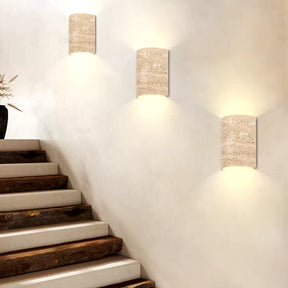 A staircase with dark wooden steps and white risers ascends on the left. Three Natural Travertine Art Deco Wall Lamps by Morsale.com illuminate the white wall on the right, casting a warm glow. A vase with dried flowers sits at the top of the stairs, enhancing the natural travertine ambiance.